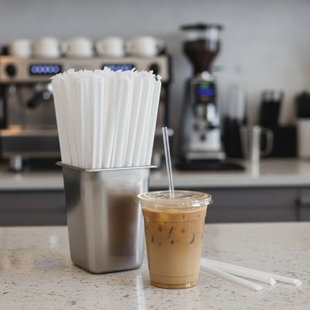 A clear iced coffee cup next to a large cluster of & Inches paper-wrapped straws standing upright in a metal holder..png
